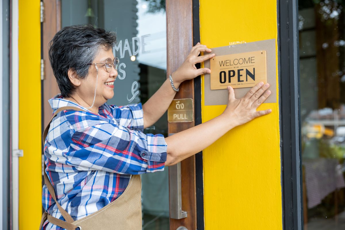 Asian senior woman puts up open sign at shop to welcome customers in the coffee shop